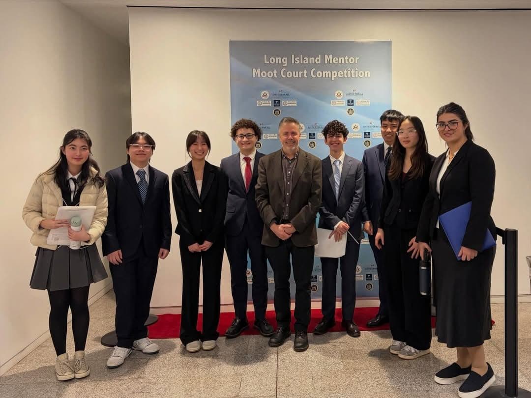 Nine students in suits and an adult standing on a red carpet in front of a blue backdrop printed Long Island Mentor Moot Court Competition.