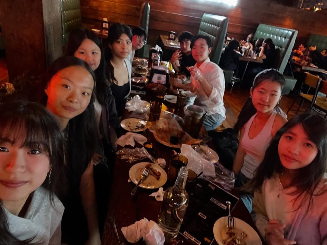 Students seated around a long wooden table in a dimly lit restaurant, looking up at the camera over shared plates.