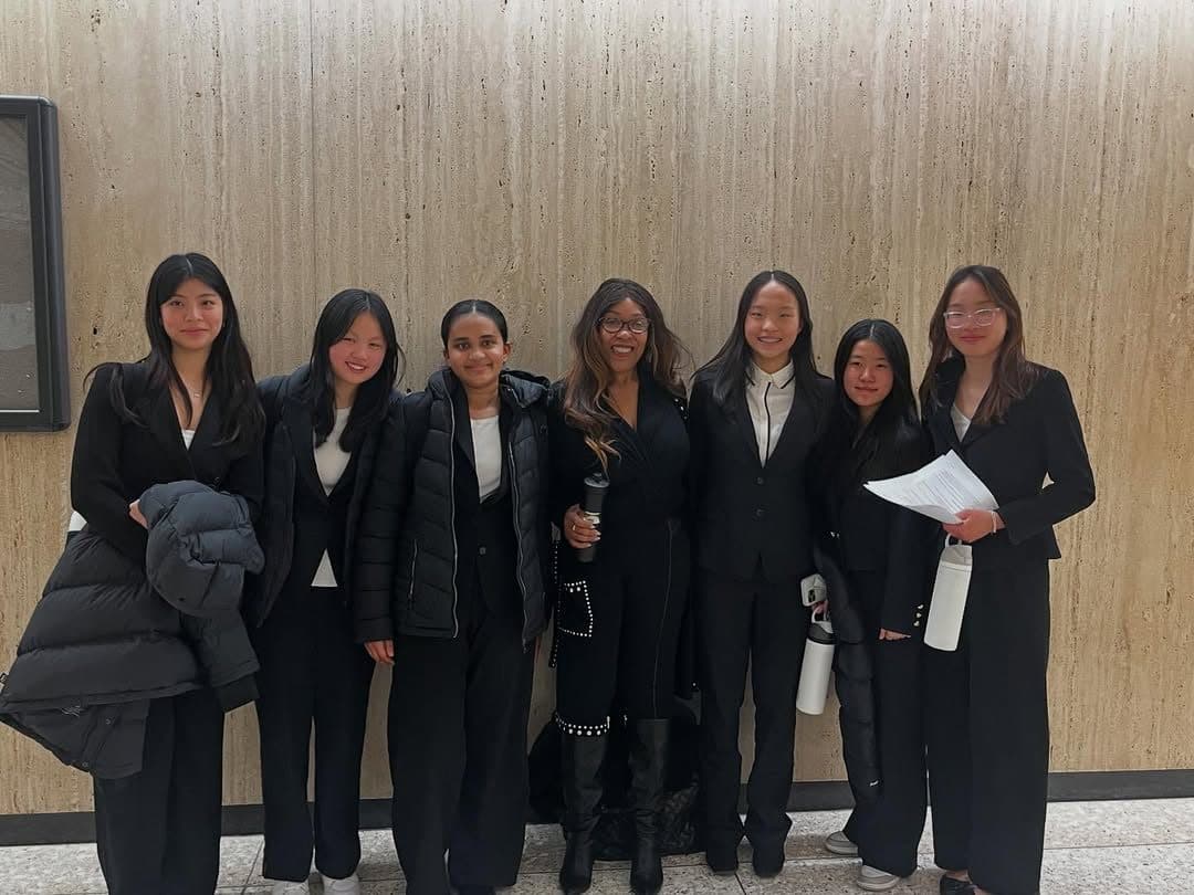 Seven people in black suits standing in a line against a pale stone wall indoors, one holding a printed document.