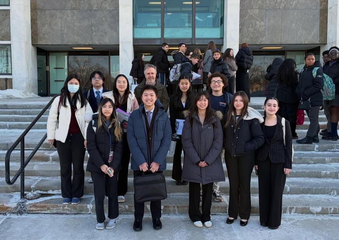 A large group of students in dark suits and winter coats on courthouse steps, some wearing face masks, with other groups gathered behind them by the entrance.